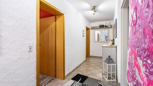 Een gang met een houten deur, een betegelde vloer, een witte wand met een structuur en een witte wand met een spiegel en een plank.