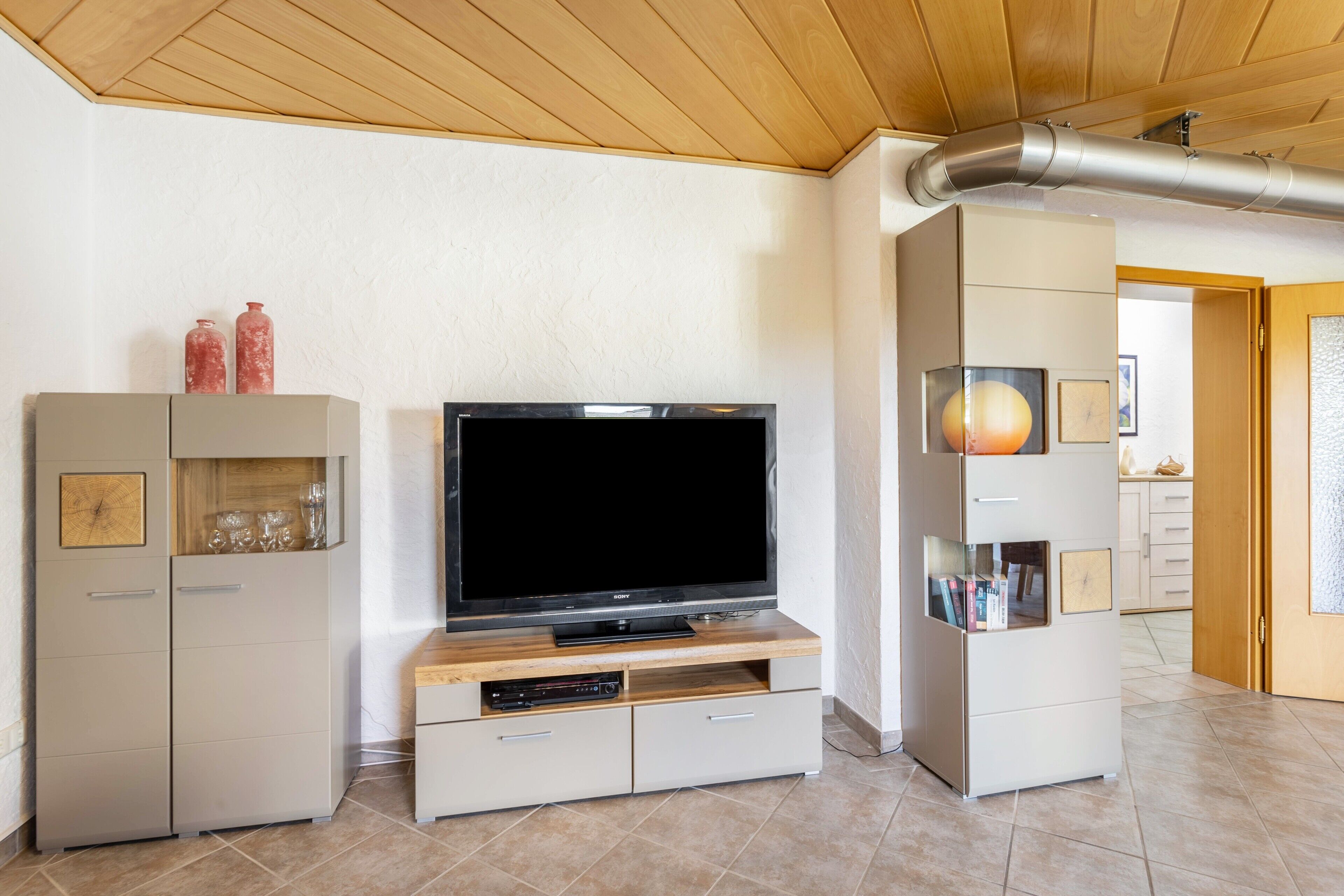 Ein modernes Wohnzimmer mit Flachbildfernseher auf einem Holzboden, einer eingebauten Vitrine mit Glastüren und einer sichtbaren Lüftungsanlage an der Decke.