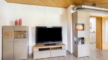 Ein modernes Wohnzimmer mit Flachbildfernseher auf einem Holzboden, einer eingebauten Vitrine mit Glastüren und einer sichtbaren Lüftungsanlage an der Decke.