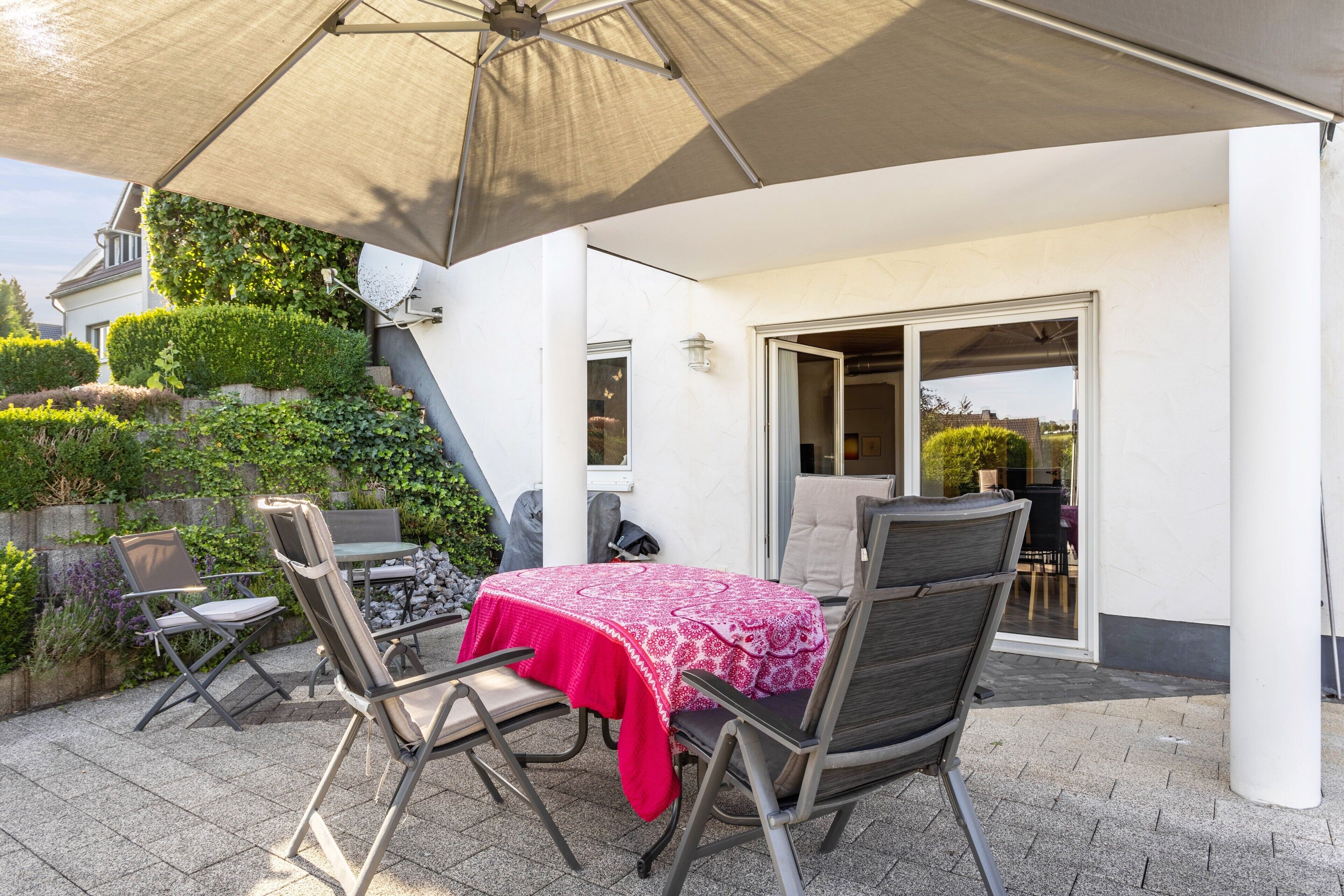 Eine Terrasse im Freien mit Tisch und Stühlen, einem großen Sonnenschirm und Blick auf ein Haus mit Garten.
