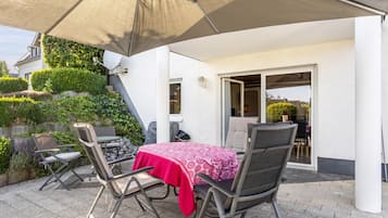 Eine Terrasse im Freien mit Tisch und Stühlen, einem großen Sonnenschirm und Blick auf ein Haus mit Garten.