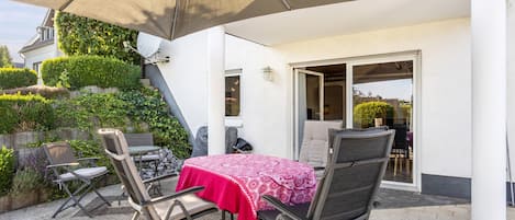An outdoor patio with a table and chairs, a large umbrella, and a view of a house with a garden.