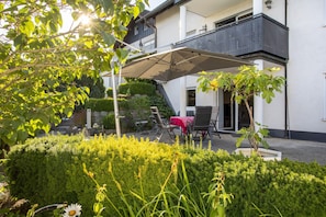 Eine Terrassenfläche mit Außenmöbeln, einem überdachten Tisch und Blick auf ein Gebäude mit einem Balkon.