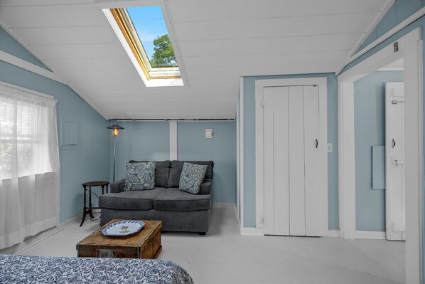 A room with a skylight, a grey sofa with patterned cushions, a small table with a plate, and a white door.