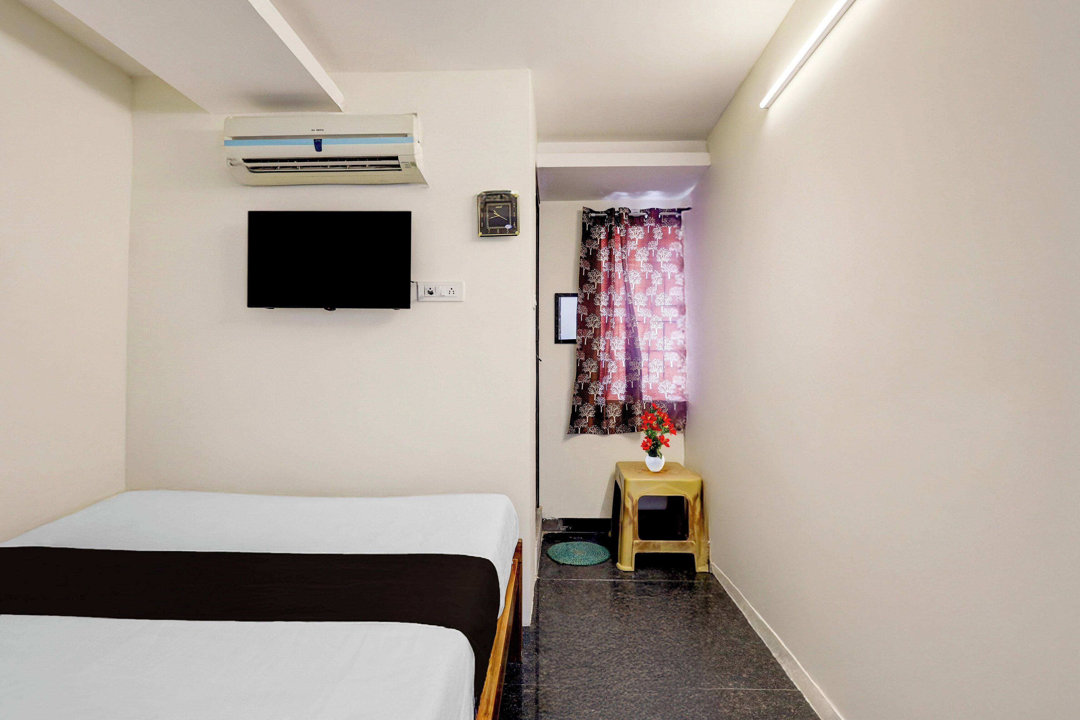 A hotel room with a bed, a TV, a small table with a vase of flowers, a clock, and a window with a curtain.
