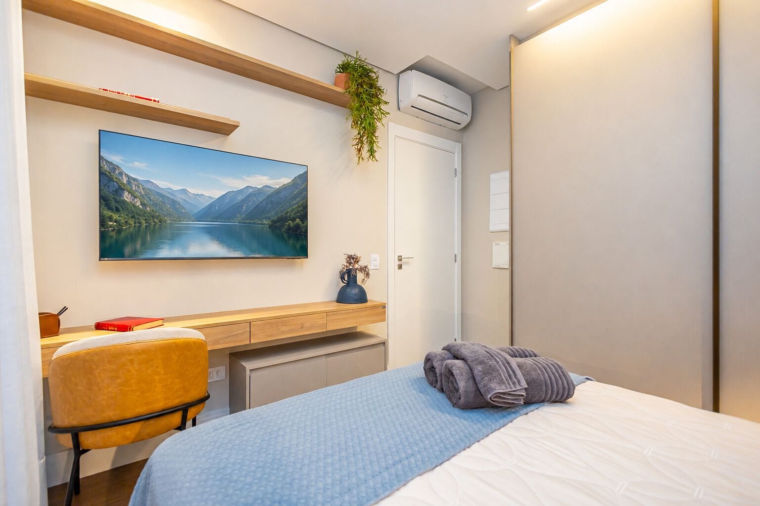A hotel room with a bed, a desk with a chair, a mounted TV, a potted plant, and an air conditioning unit.