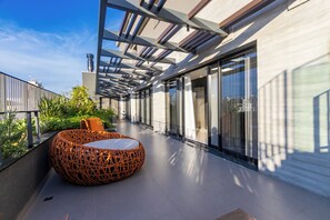 Un área de estar exterior moderna con una mesa y sillas de mimbre redonda, ventanas amplias y cielo despejado.