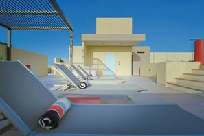 Un área de piscina en la azotea con sillas de descanso, una escalera y un edificio con una puerta azul claro.