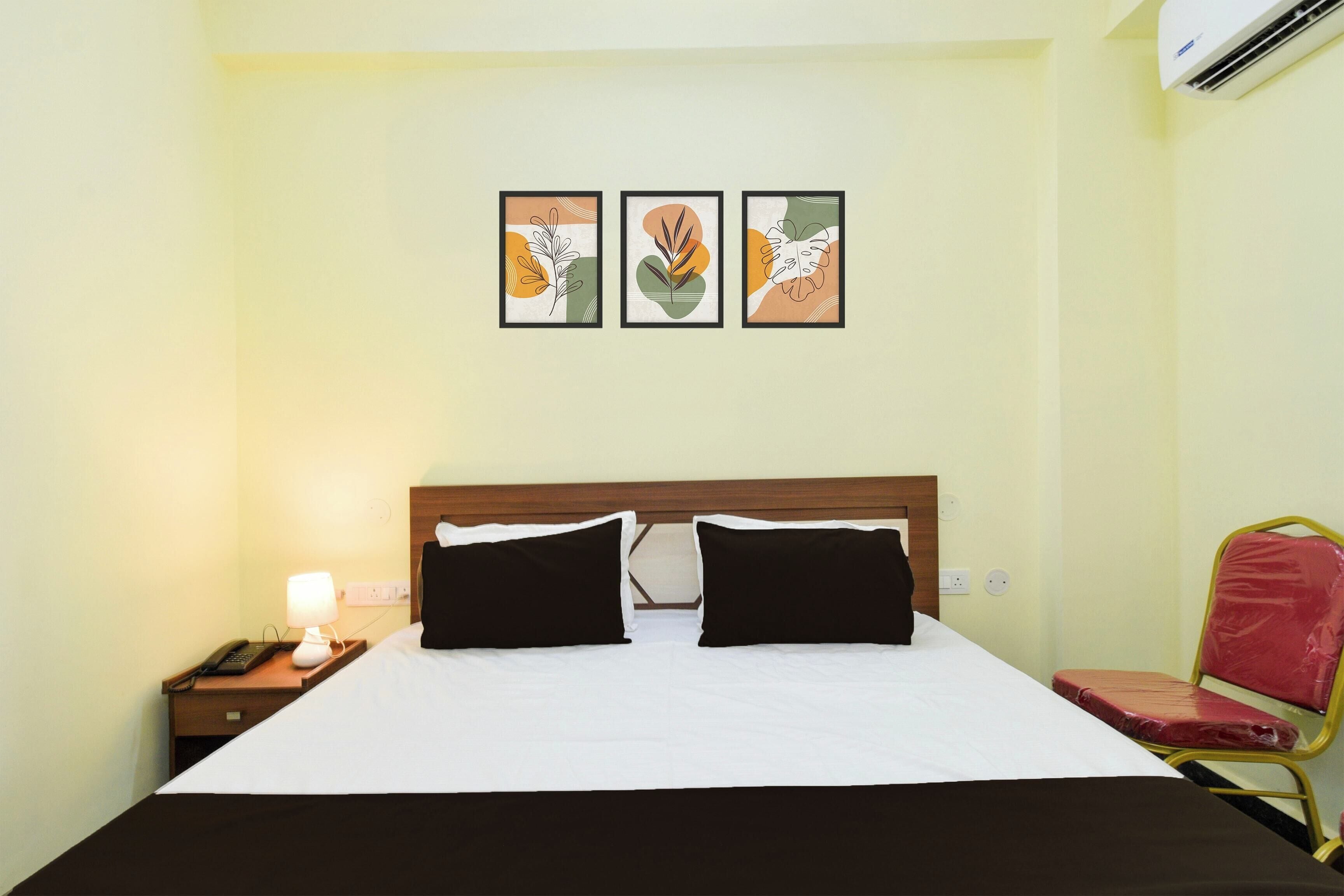 A hotel room with a bed, two black pillows, a nightstand with a lamp, a phone, and a wall-mounted air conditioner.