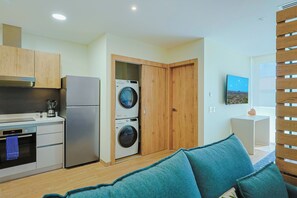 Cocina compacta con refrigerador, estufa y lavadora, una puerta de madera y un pequeño rincón de descanso con cojines.
