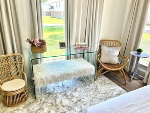 Ein Zimmer mit Glastisch, zwei Korbstühlen, einem flauschigen Teppich und Blick auf ein Haus durch transparente Vorhänge.