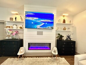 Ein Wohnzimmer mit einem an einer weißen Wand montierten Flachbildfernseher, einem Kamin darunter und zwei Beistelltischen mit Dekorationselementen.