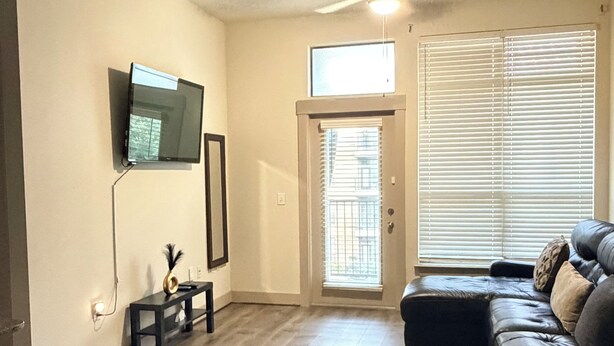 Uma sala de estar com televisão de tela plana na parede, um ventilador de teto, um sofá de couro preto e piso de madeira.