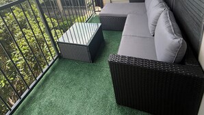 Uma varanda com sofá, mesa, grade e vista para um edifício e vegetação.