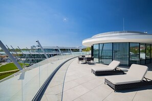 Un espace de détente extérieur moderne, doté de sièges et d’une balustrade en verre donnant sur un port de plaisance.