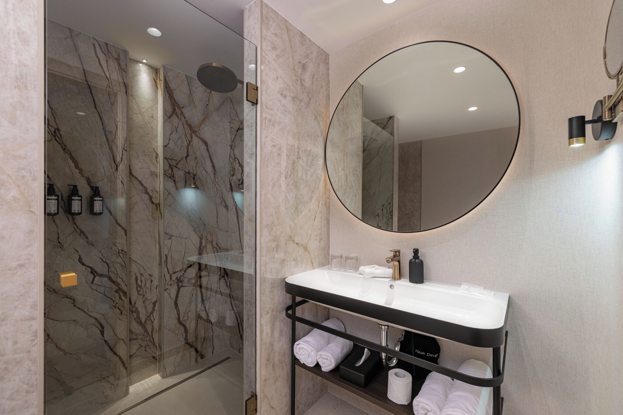 Een moderne badkamer met een marmeren douche, een ronde spiegel, een witte wastafel en een handdoekrek.