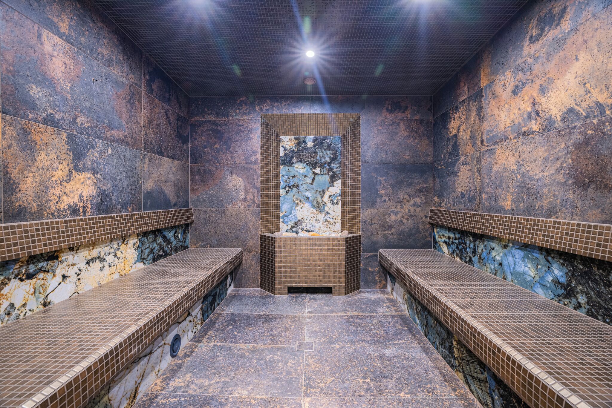 Un sauna con dos bancos, una pared central con una obra de arte texturizada y suelo de baldosas.
