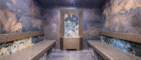 Un sauna con dos bancos, una pared central con una obra de arte texturizada y suelo de baldosas.