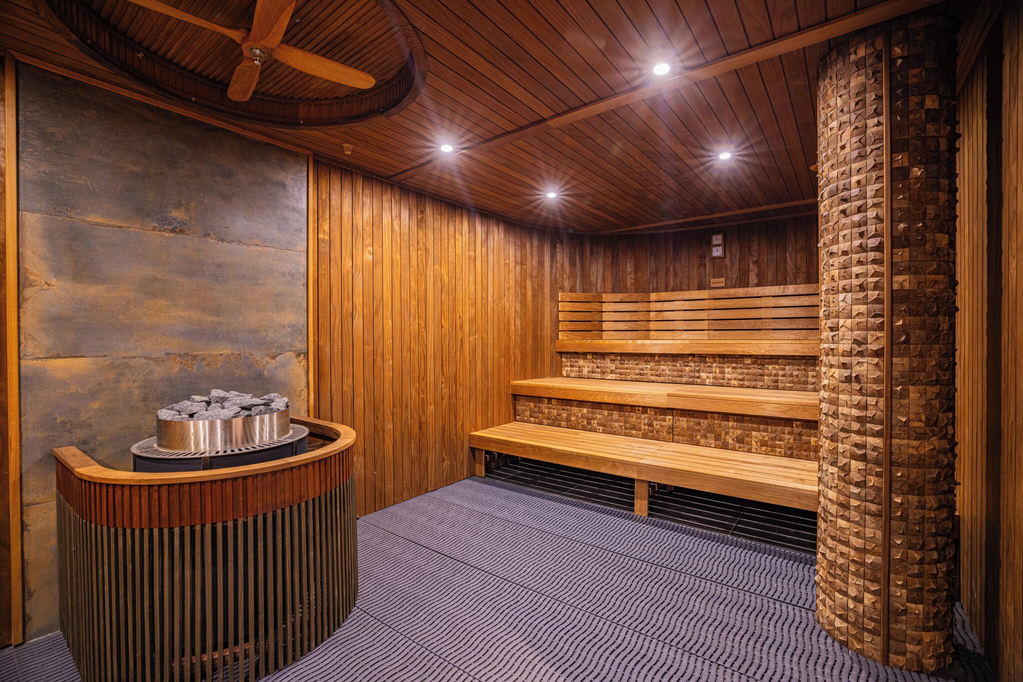 A sauna room with wooden benches, a stone wall, and a wooden ceiling with a fan.