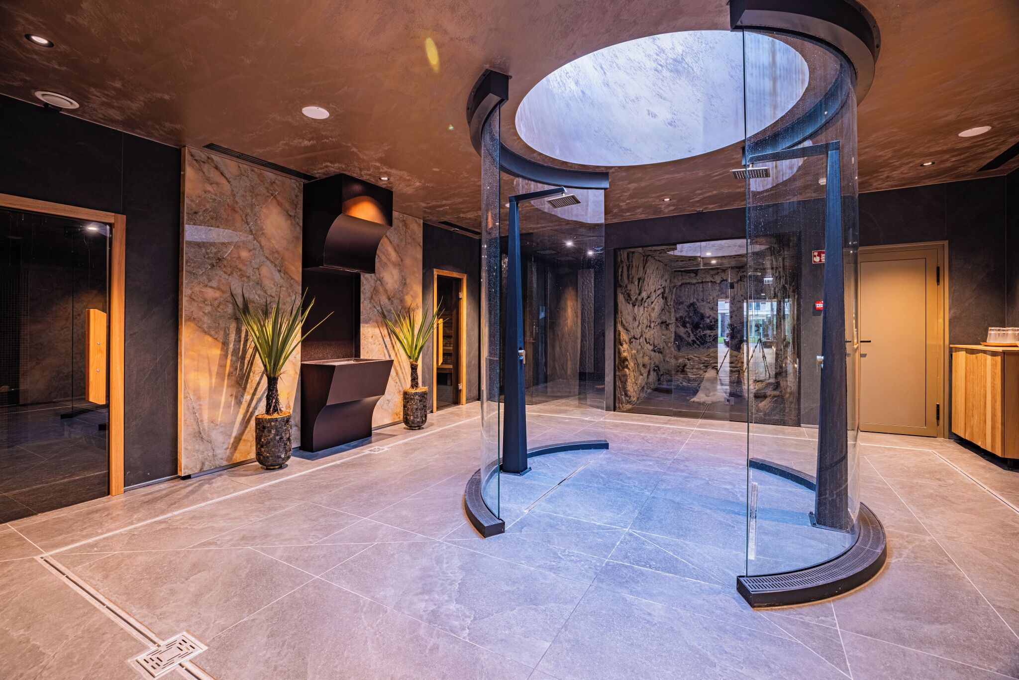 A modern lobby with a circular glass elevator, marble flooring, and a reception desk.