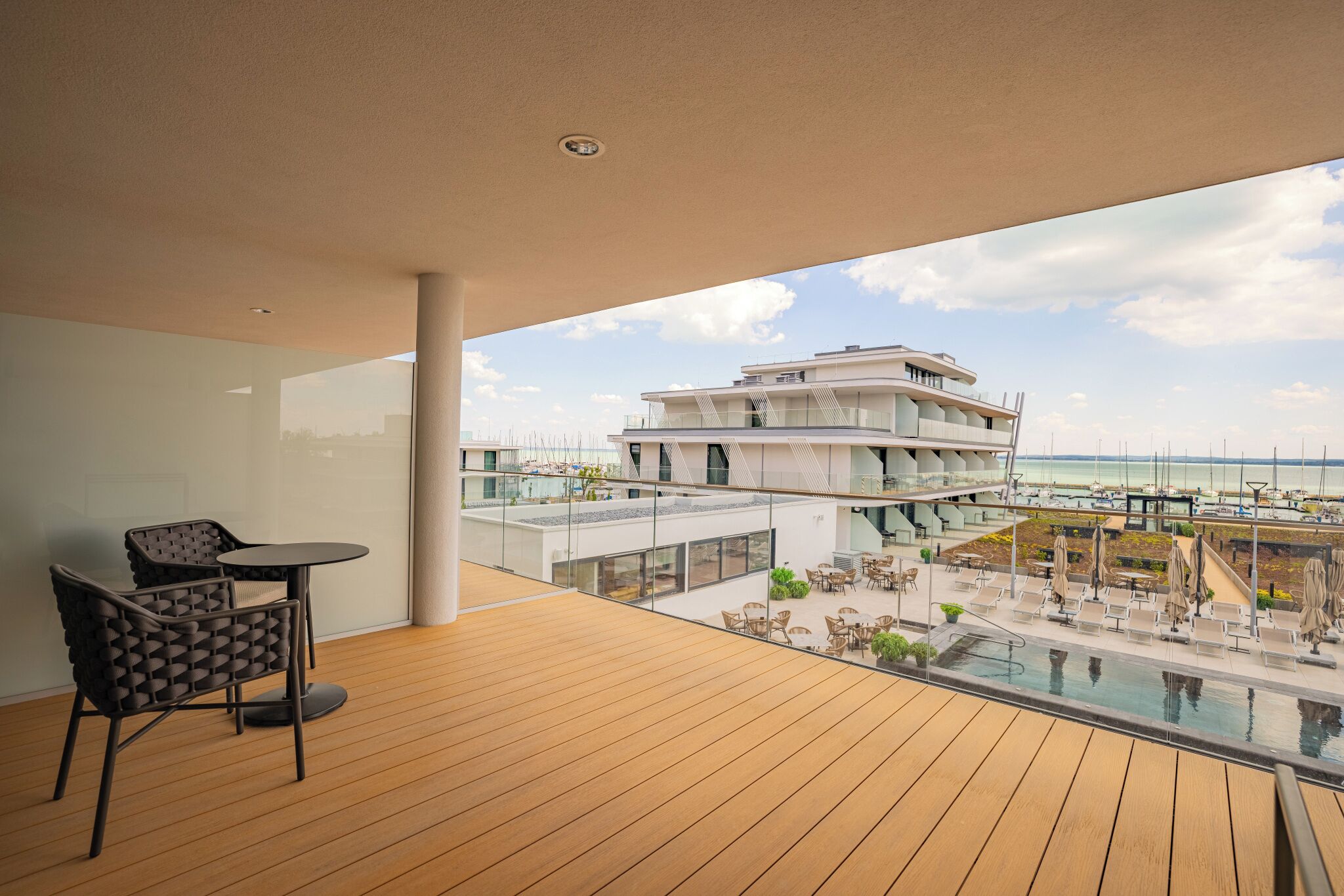 Un balcón de hotel moderno con suelo de madera, barandilla de vidrio y vista a un puerto deportivo.