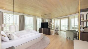 Una habitación de hotel moderna con una cama grande, televisión, zona de estar y ventanas amplias.