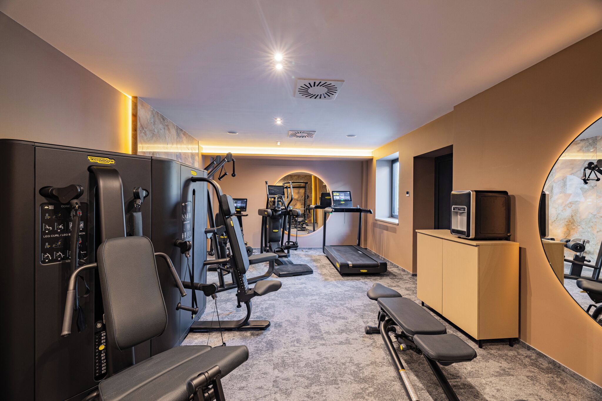 Un gimnasio con máquinas de cardio, equipo de pesas y un espejo grande.