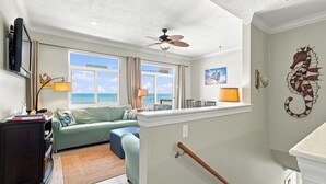 Ein Zimmer mit Deckenventilator, Blick auf das Meer durch große Fenster, einer Wanddekoration mit einem Seepferdchen und einer Holzbank.
