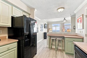 A kitchen with a black refrigerator, green cabinets, a bar area with stools, and a view through the window. - Gulf Front Condo  at Seahorse Condos | Dog Friendly | Sleeps 9 (Pensacola Beach)