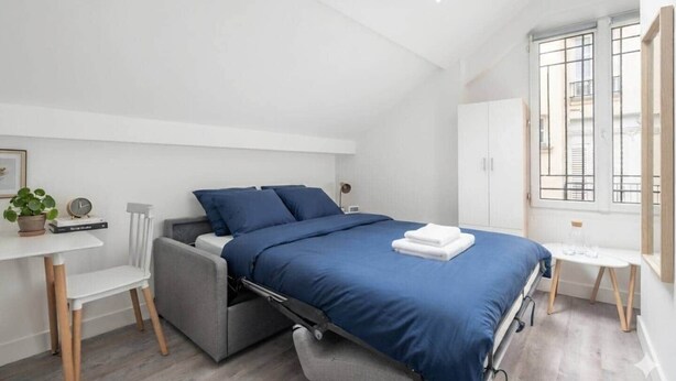 A modern bedroom with a skylight, a bed with blue bedding, a small table with a plant, and a window with bars. - Charming Studio Near the Eiffel Tower (Paris)