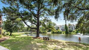 A lakeside park with a bench, a pond, and buildings in the background. - Lagoon View Villa Near Beach and Tennis (Hilton Head Island)