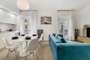 Een moderne woonkamer met een blauwgroene bank, een eetgedeelte met een witte tafel en stoelen, en een keuken met witte kastjes.