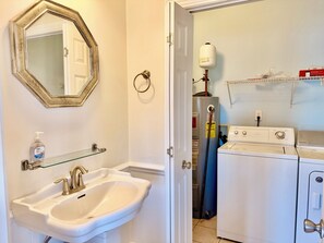 A bathroom with a sink, mirror, and a door leading to a laundry area with a washing machine. - Old Bay Watch: 3BR 2.5BA, Walk to Beach, Boat Parking, Sleeps 6 (Oak Island)