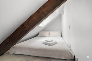 Ein kleiner Raum mit weißer, schräger Decke, ausgestattet mit einem Bett, Kissen und einer ordentlich gefalteten Decke.
