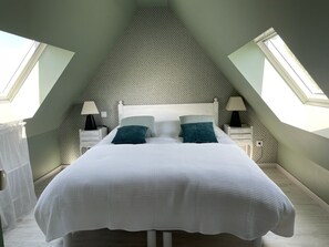 Une chambre avec un plafond en pente, un lit recouvert d’une literie blanche, deux tables de chevet avec des lampes et une fenêtre sur le côté gauche.