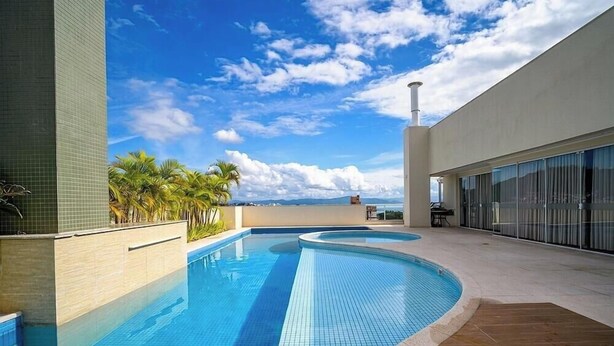 A swimming pool with a curved edge, surrounded by a tiled deck and palm trees, with a clear sky above. - Apt. in Condo. w/ Pool & Garage Pea0410 (Florianópolis)