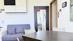 Une pièce avec une table en bois foncé, un canapé gris, des chaises blanches et un mur blanc orné d’un tableau encadré.