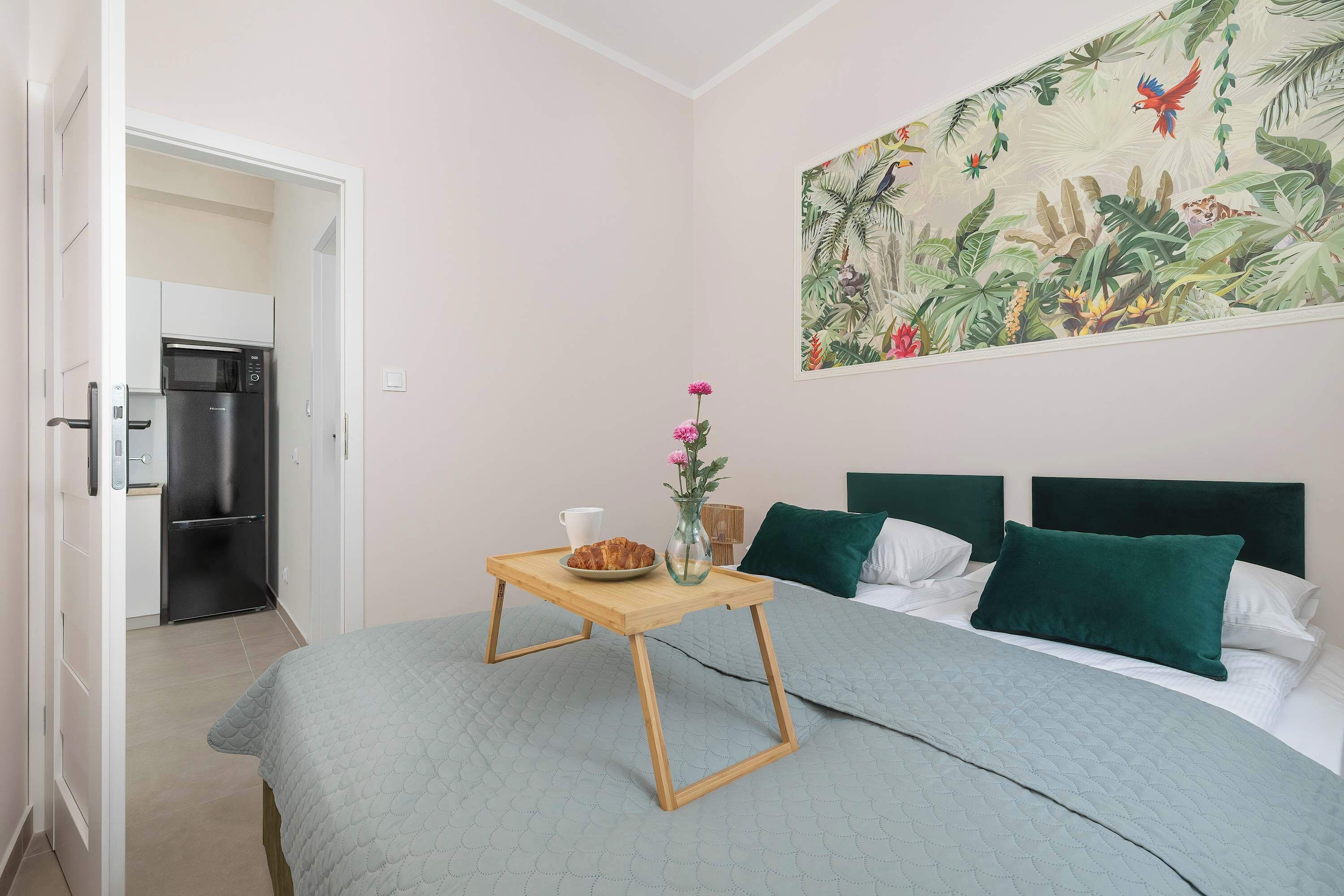 Une chambre à coucher avec un lit, une table de chevet en bois sur laquelle se trouvent une tasse et un vase avec une fleur, une tête de lit verte et un tableau mural.
