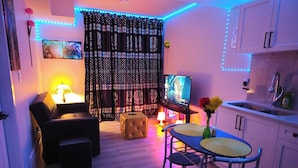 Olohuoneessa on sohva, rahi ja jalustalla oleva televisio. Siellä on myös ruokailutila, jossa on pöytä ja tuolit, sekä keittiö, jossa on kaappeja ja pesuallas.