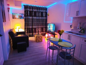 Uma sala de estar com sofá, banquinho e televisão em um móvel. Há uma área de jantar com mesa e cadeiras, e uma cozinha com armários e pia.
