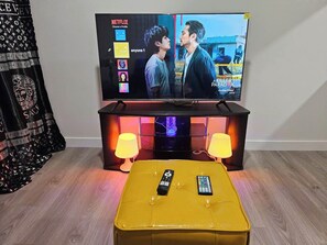 Uma televisão sobre um suporte com interface Netflix, dois controles remotos sobre um banquinho amarelo, e uma tapeçaria estampada na parede.