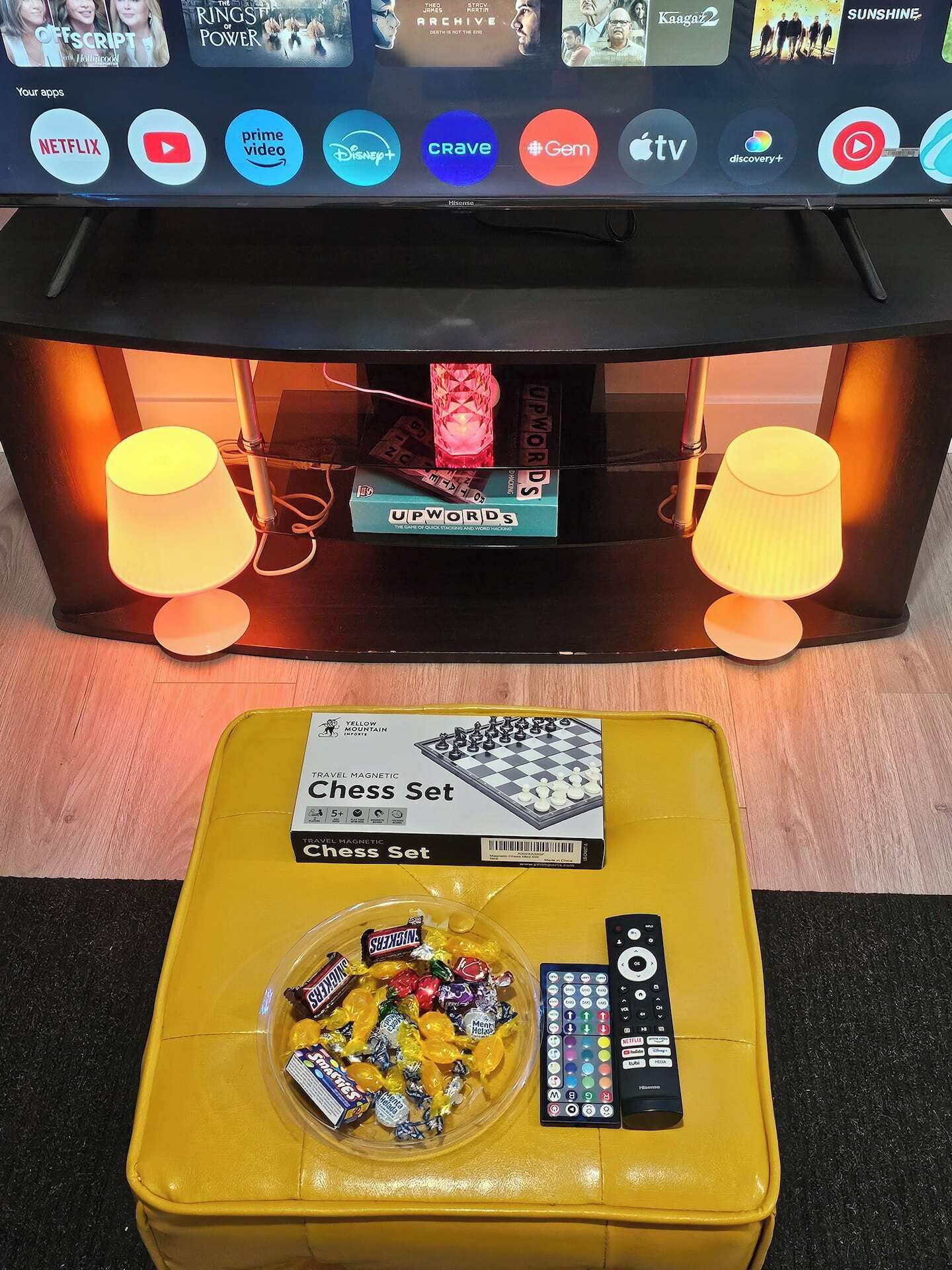 A television with a game box labeled 'UPWORDS' and a yellow ottoman with a chess set, chocolates, and a remote control.