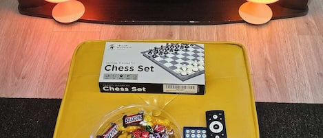 En tv med ett spel som heter ”UPWORDS” och en gul ottoman med ett schackset, choklad och en fjärrkontroll.