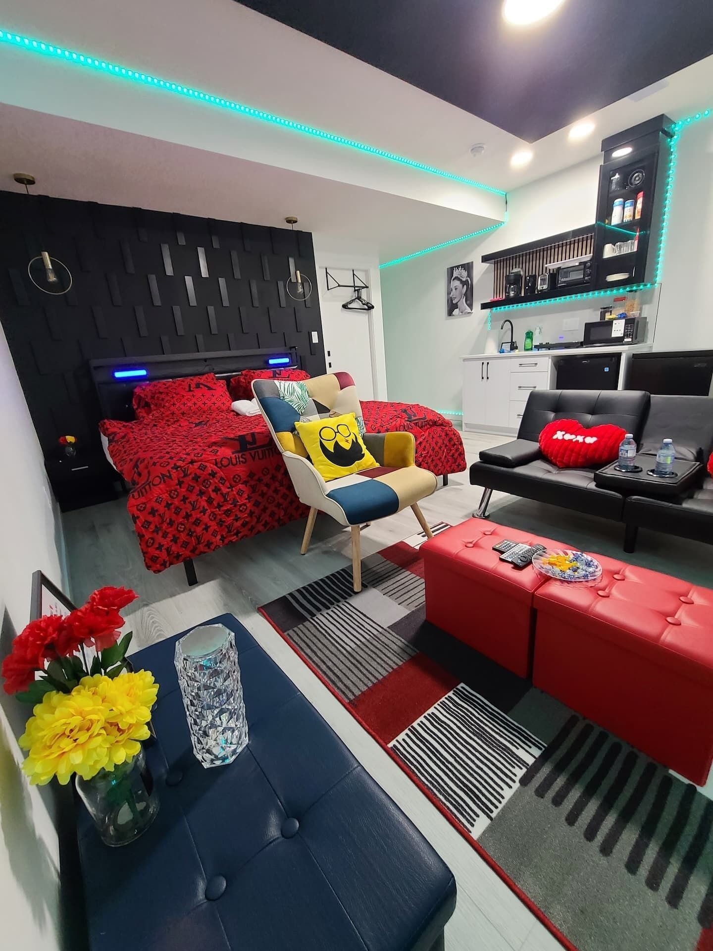 A modern living room with a sofa, ottoman, and a bed with a red patterned bedspread.