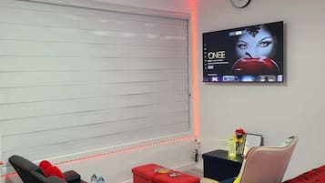 Una habitación de hotel moderna con televisión de pantalla plana en la pared, un sofá, un reposapiés rojo y una cama con colcha estampada en rojo.