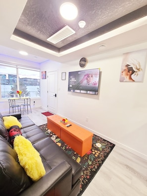 A modern living room with a black leather sofa, an orange ottoman, and a flat-screen TV mounted on the wall. - SwitHome Properties Cher Cozy Suite (Pond View), Near YEG Airport. (Edmonton)