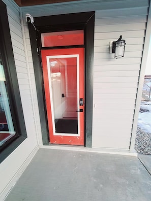 A red door with a peephole and a keypad lock, a small outdoor light fixture, and a concrete step leading to the door. - SwitHome Properties Cher Cozy Suite (Pond View), Near YEG Airport. (Edmonton)