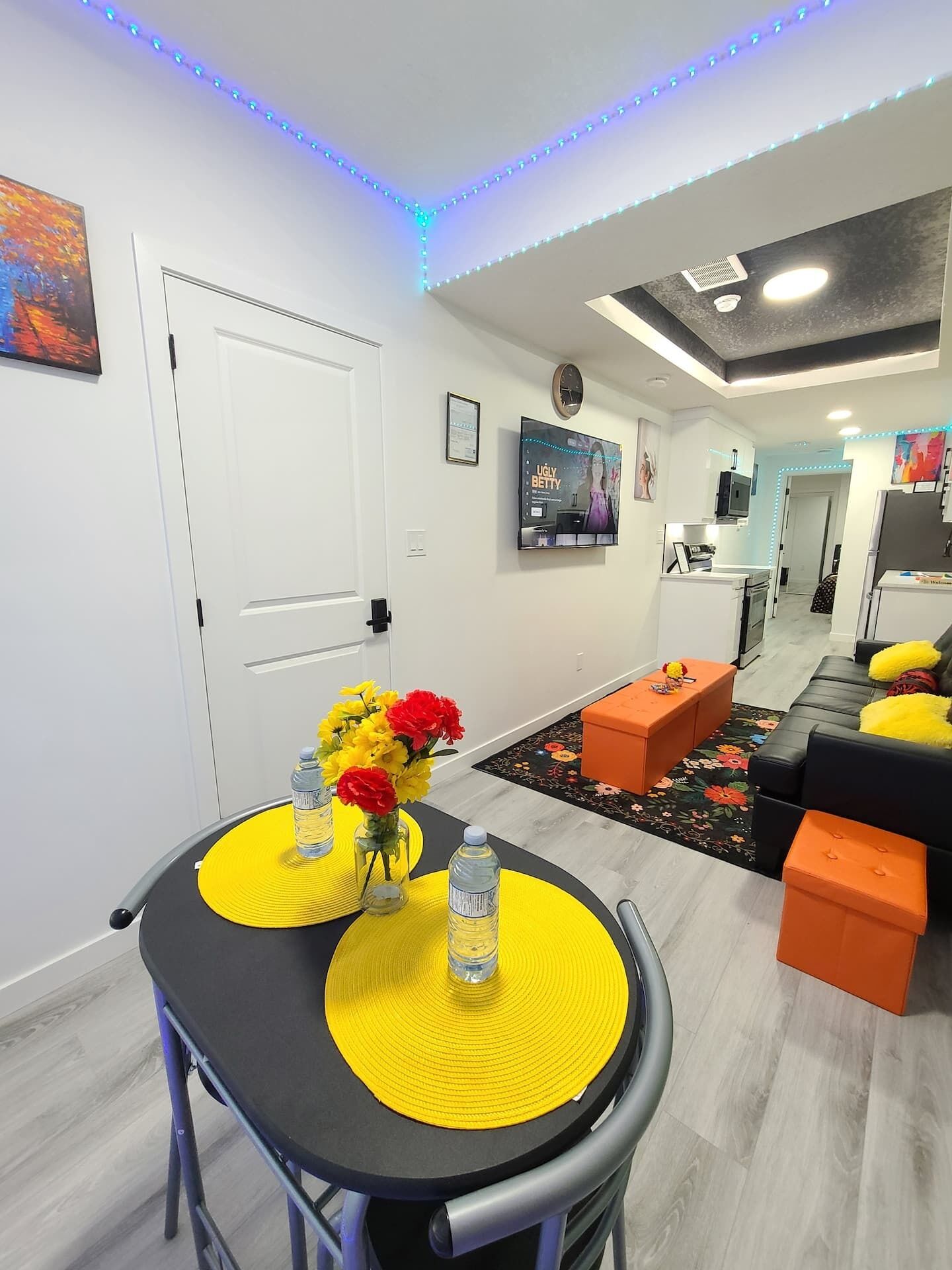 A modern living room with a dining area, featuring a sofa, ottomans, a TV, and a painting on the wall.