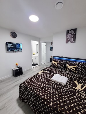 A modern bedroom with a large bed, a flat-screen TV, a bathroom visible through an open door, and a ceiling light. - SwitHome Properties Cher Cozy Suite (Pond View), Near YEG Airport. (Edmonton)