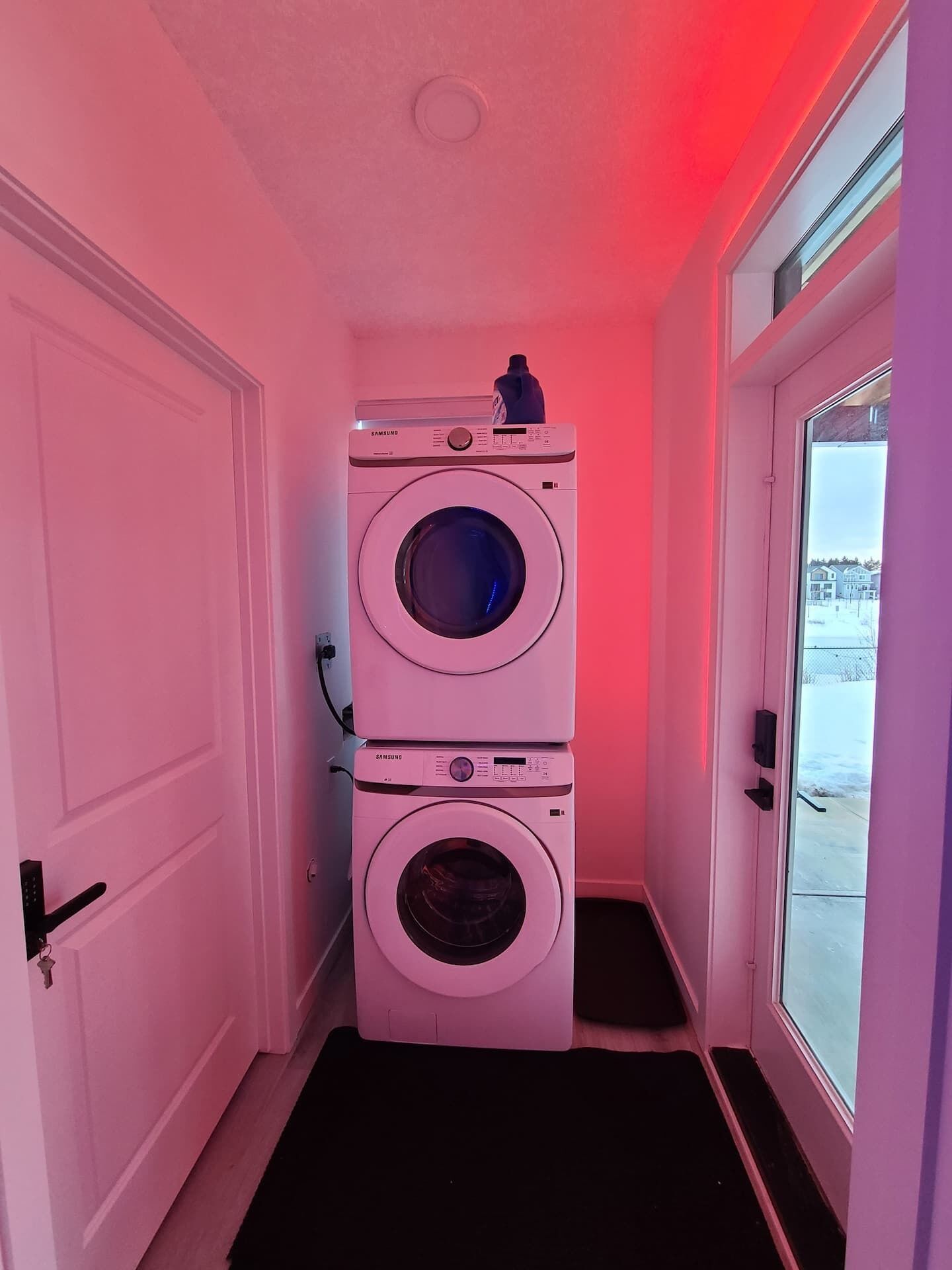 A laundry room with two stacked washing machines, a door with a key lock, and a window with a view of a snowy landscape.